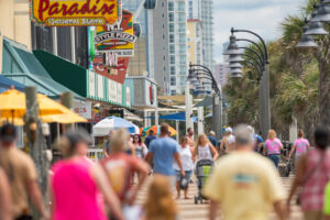 A vibrant, bustling scene on the Myrtle Beach Boardwalk, featuring crowds of vacationers strolling past colorful storefronts, gift shops, and outdoor eateries under a row of classic streetlamps. This high-energy coastal setting highlights the world-class entertainment and oceanfront excitement available to guests booking with Casago Myrtle Beach.