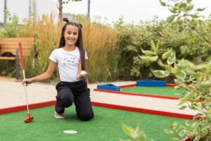 A smiling young girl holding a golf ball and putter while kneeling on a vibrant green miniature golf course. This cheerful scene highlights the numerous family-friendly attractions and classic vacation activities available to guests staying with Casago Myrtle Beach.
